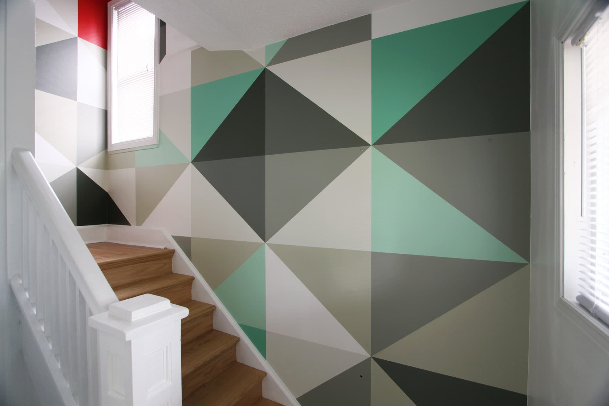 Triangulated mural in mint, sage, and graphite climbing a residential stairwell, with a single red triangle at the window.